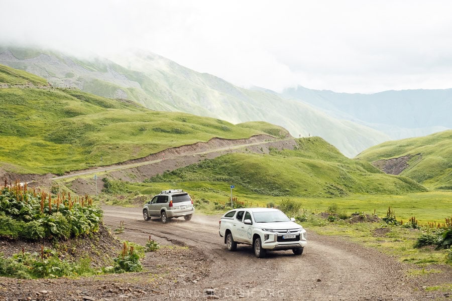 Two cars on the road to Khevsureti, surrounded by green mountains.