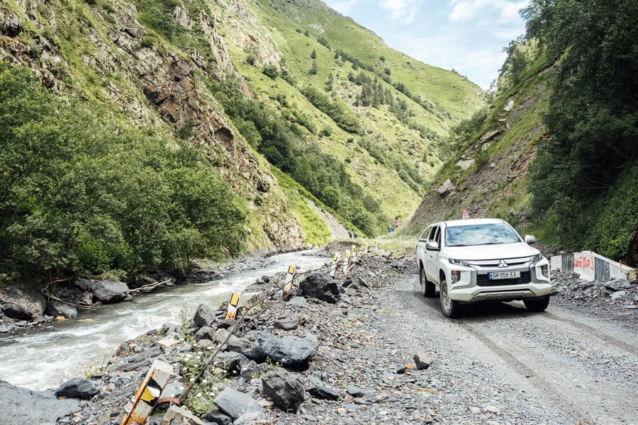 A white car driving on a gravel road alongside a river in Khevsureti, with a safety barricade falling away into the gorge.