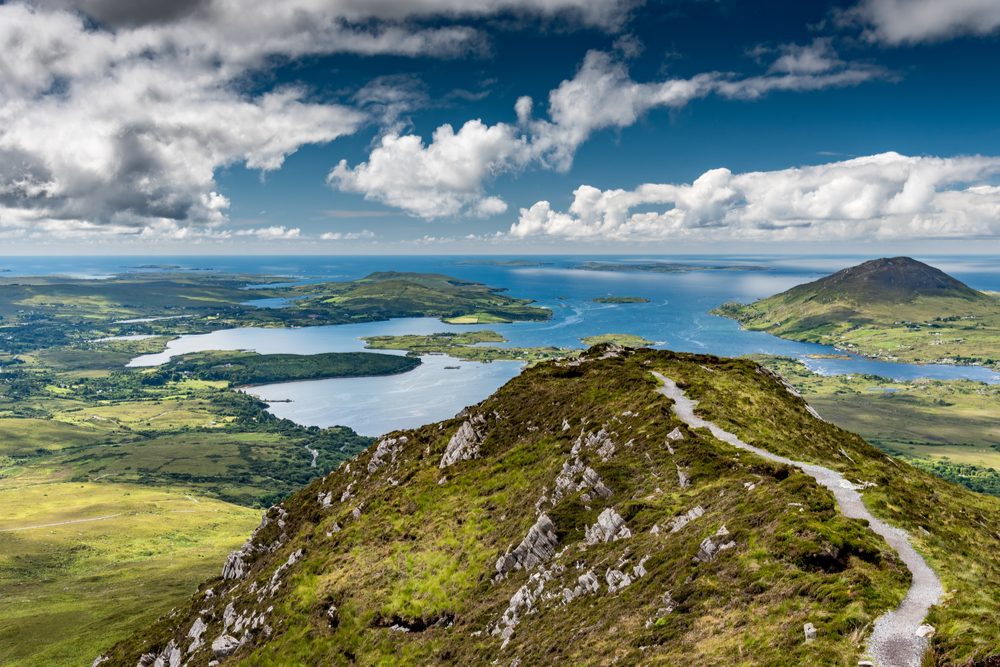 The hiking trail at the top of Diamond Hill in Connemara National Park, Ireland. Behind, the sun plays with the clouds reflected in the sea. - Image