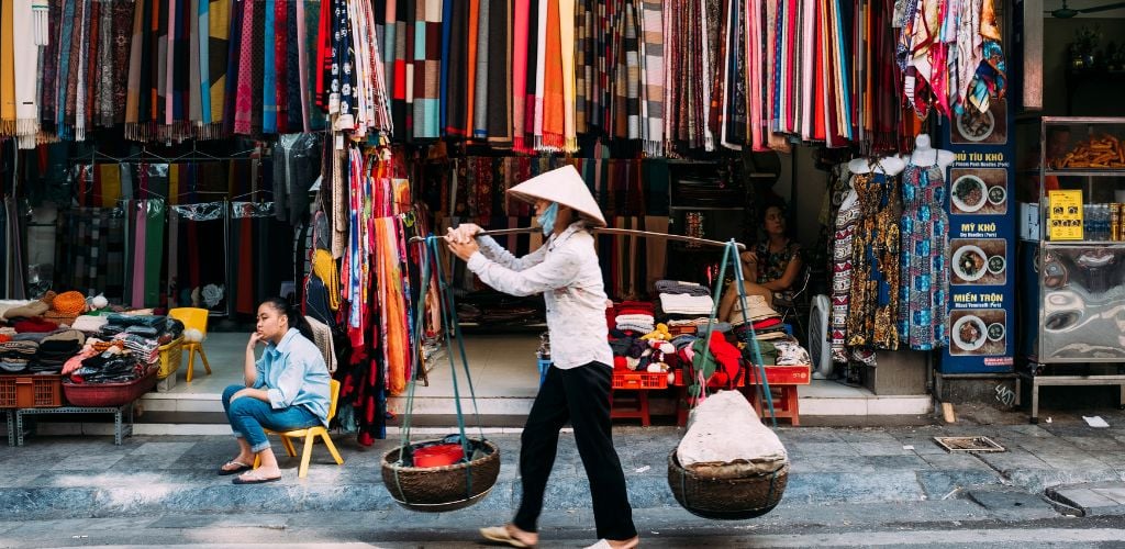 store owner sitting on low stall in front of colourful hanging materials whilst another street vendor walks, Hanoi 