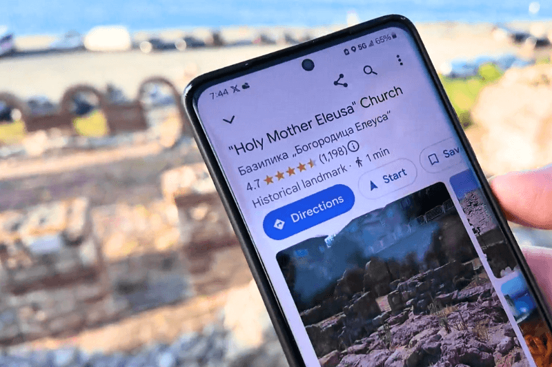 A hand holds a phone with the Google map listing open for Holy Mother Eeusa Church in Nessebar. The ruins are slightly visible in the faded background.