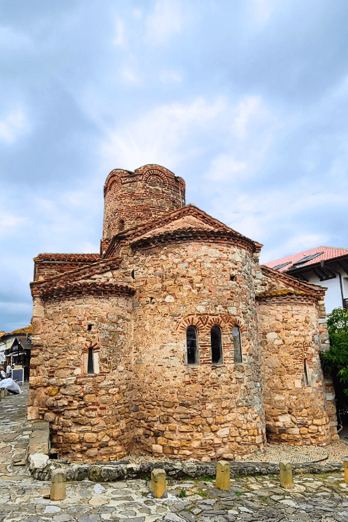 Church of St John the Baptist is one of the oldest looking in Nessebar. Rough sandstone colored bricks make up the three rounded alcoves of the outside of the church. A small girl in pink with a short brown bob stands in front of the Church.