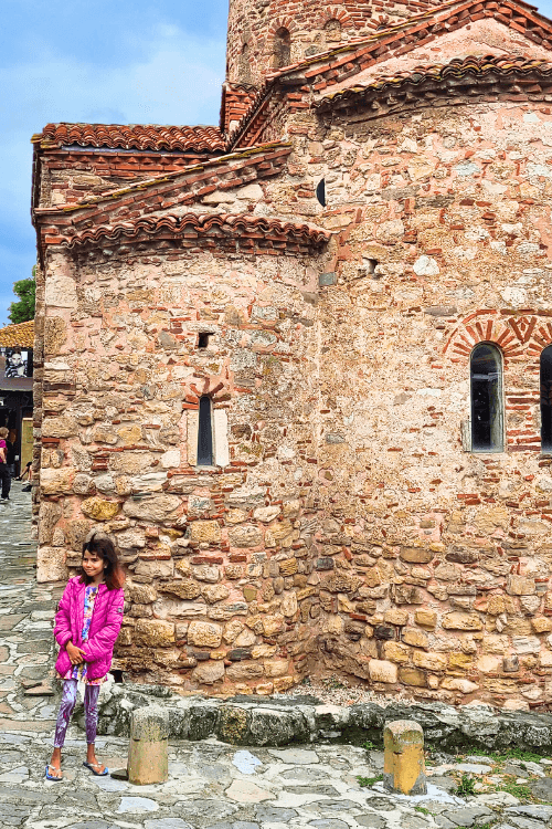 Church of St John the Baptist is one of the oldest looking in Nessebar. Rough sandstone colored bricks make up the three rounded alcoves of the outside of the church. A small girl in pink with a short brown bob stands in front of the Church.