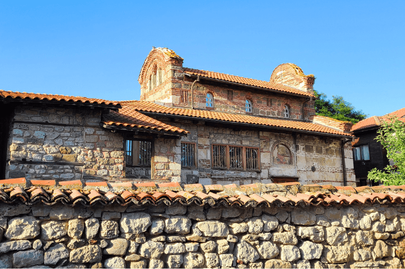 The ancient church of St Stephen in Nessebar features a tall arched facade on either end and rough ancient stone. A stone wall with typical orange roof tiles sits in the foreground.