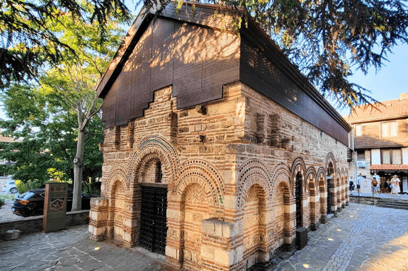 The Church of Saint Paraskeva stands mostly in tact, but with a modern metal roof. Trees filter the sun on the front of the Church.