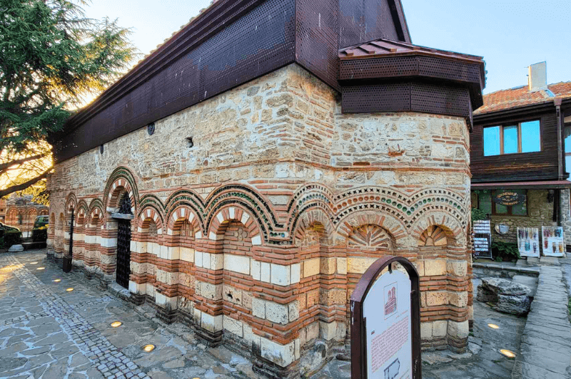 Side wide angle shot of the Church of Saint paraskeva in Nessebar Bulgaria which features long walls of white and red arches with stacked stone on top. The original roof is gone and has been replaced with a bronze colored metal one.