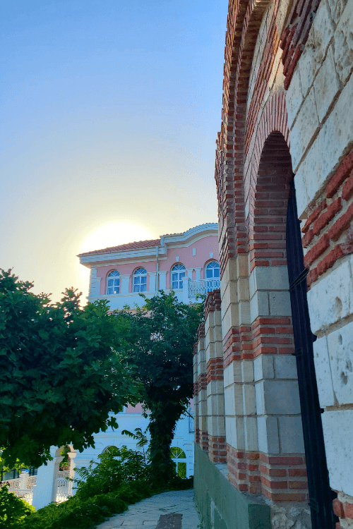 The white and red layered ancient brick of St John Alitourgetos Church in the foreground with a tall pink building in the background and green trees in between.