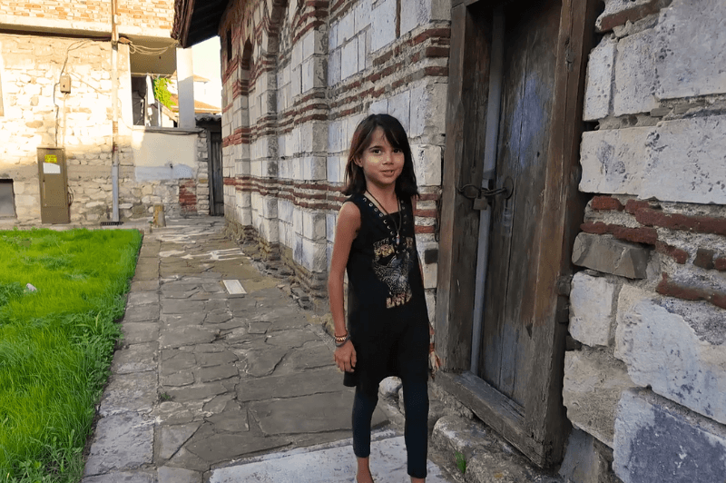 A small girl with short dark hair stands outside a very small door into the side of a typical Nessebar church in white and red stone: St Todor Church.