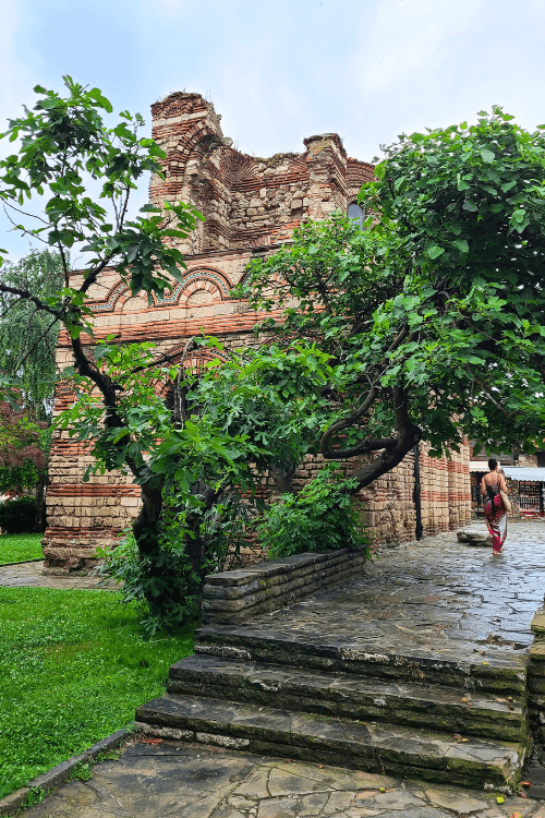 Trees shroud the distant Christ the Pantocrator Church in Nessebar Bulgaria. A stone sidewalk and stairs lead up to it.