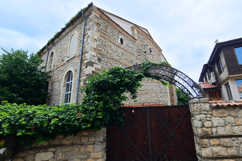 The tall three storey stone church of Dormtion of Theotokus in Nessebar appears over top of a stone wall covered in vines with a large black gate.