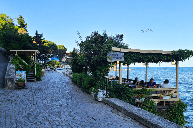 A stone street close to the waterfront on the south side of Nessebar peninsula is lined with restaurants beside the sea. The sky is cloudless and blue, with gulls flying overhead.