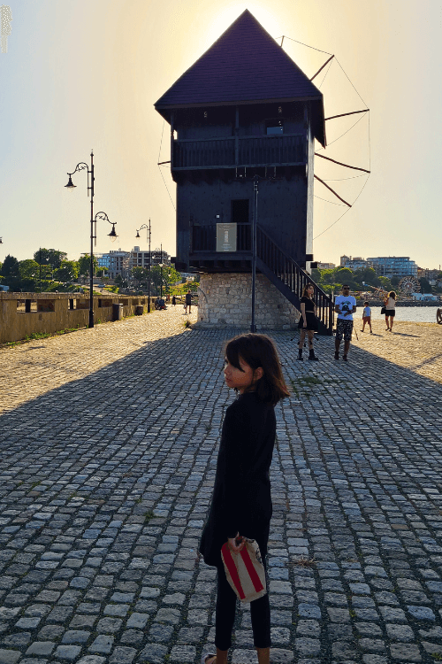 A girl with short brown hair stands on a cobbled street in the long shadow of an ancient wooden windmill in Nessebar Bulgaria near sunset.