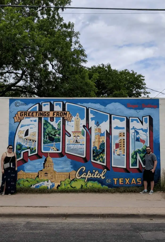 Riana and Colin standing in front of a mural in Austin, Texas that reads "Greetings from Austin, Capitol of Texas"