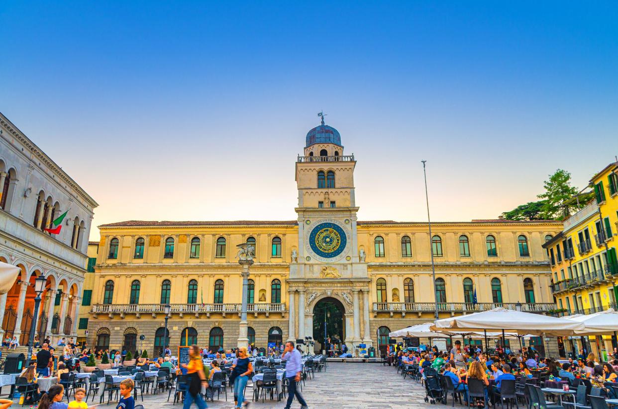 Padua, Italy, September 12, 2019: Torre dell'Orologio clock tower with astronomical clock and restaurant tables in the Piazza Plaza Dei Signori square, twilight evening view, Veneto Region