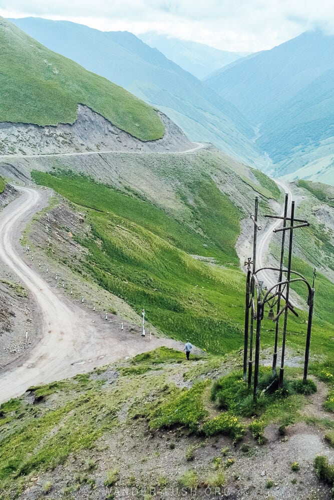 A metal cross mounted on a hill with a man walking down towards the winding road at the Datvisjvari Pass in Georgia.