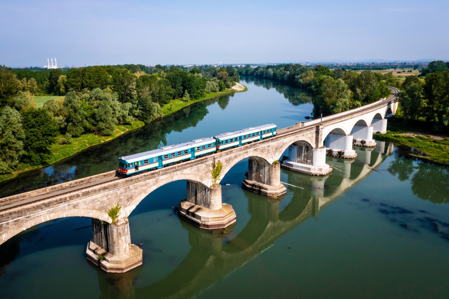 Aerial view of vintage turquoise and white train traveling across river on a bridge with multiple archways 