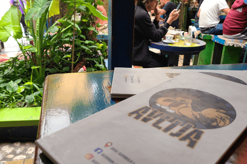 The menu for Avlija sits in the foreground on a wooden table. In the background there are lots of plants and people sitting at brightly painted tables.