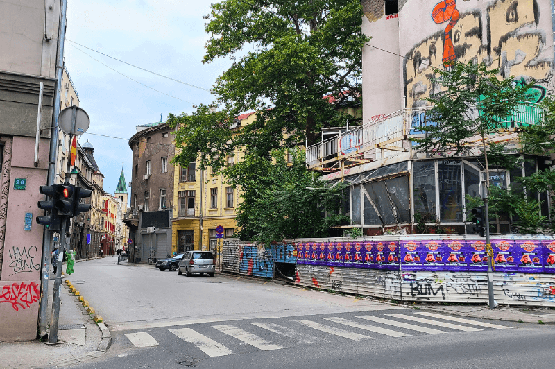 A delapidated building on the right side of a street corner in Sarajevo behind a fence covered in posters. At the end of the long street in the distance you can see the spire of a chapel.