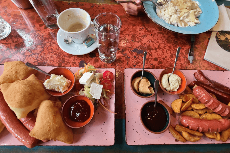 a breakfast spread of deep fried bread, sausages, jam, and french fries on two pink plates over a red table at Avlija Restaurant in Sarajevo.