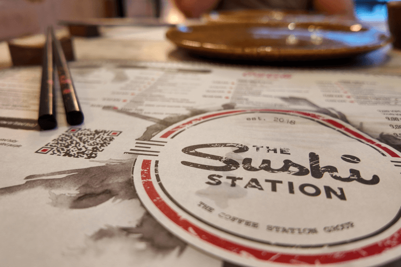 A paper menu reads "sushi station" with a pair of chopsticks resting in the background.