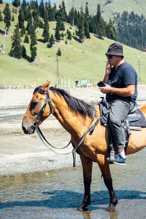 A man smoking a cigarette and looking at his mobile phone whilst sitting on a horse in Bakhmaro.