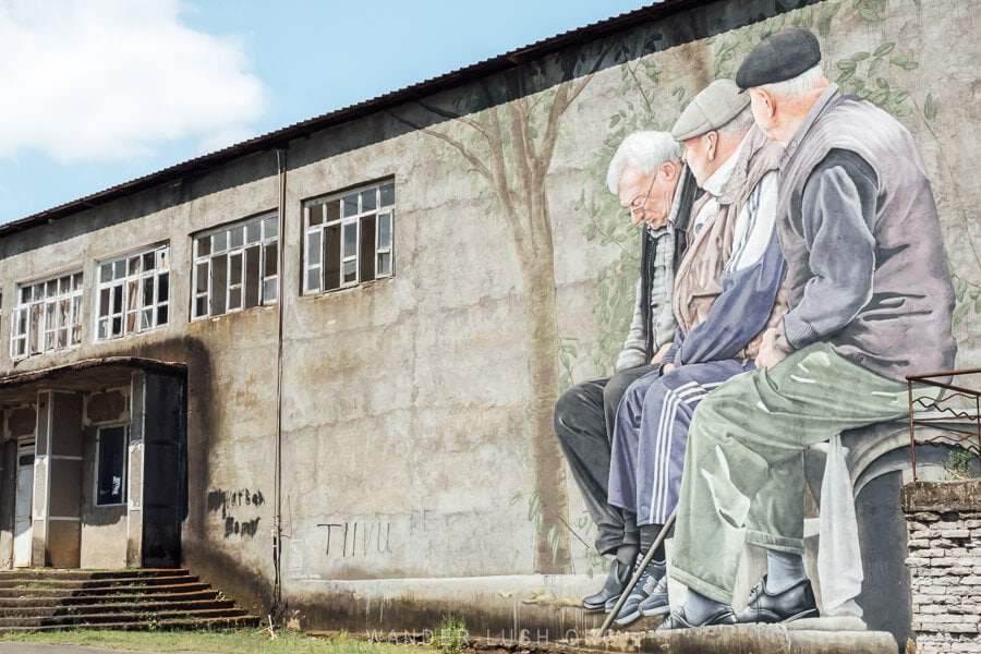 A large mural depicting three old men on the side of a concrete building in a village in Guria, Georgia.