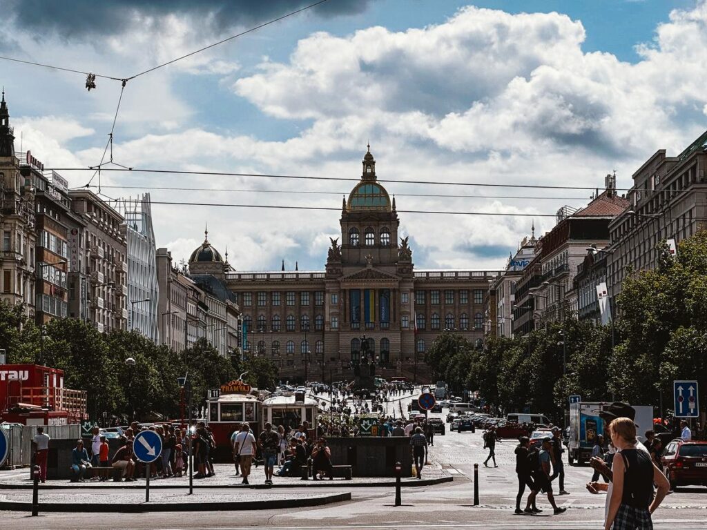 A lively street view with the National Museum in Prague, Czech Republic, featuring pedestrians, trams, and historic architecture under a cloud-filled sky.