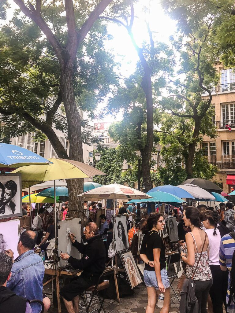 Street artists painting portraits under colorful umbrellas at Place du Tertre in Montmartre, Paris, with crowds of people enjoying the lively artistic ambiance.