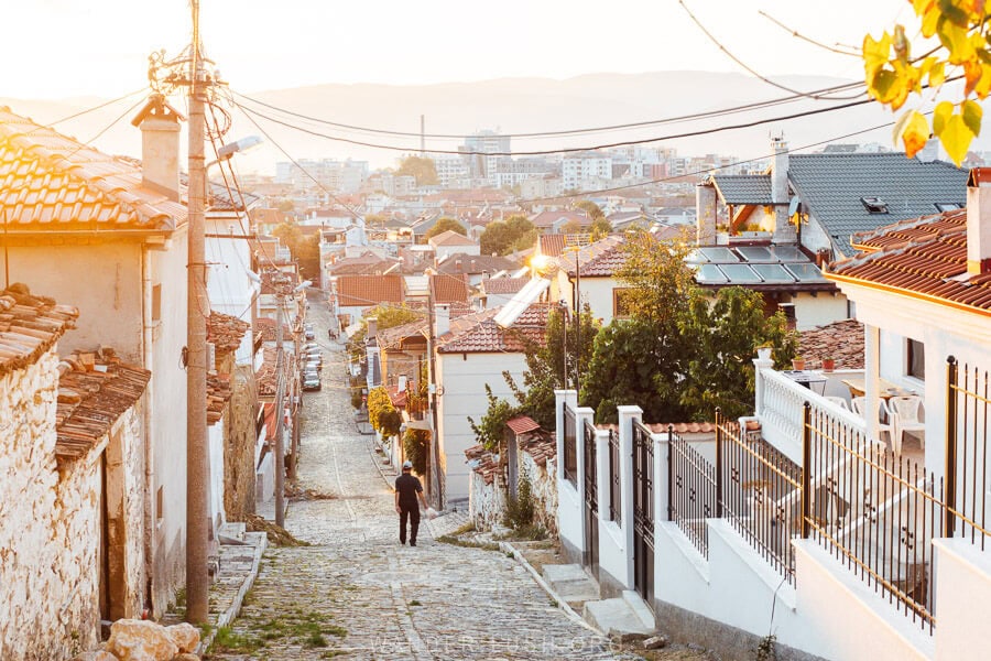 A man ambles down a cobble street between houses in the city of Korca, Albania at sunset.