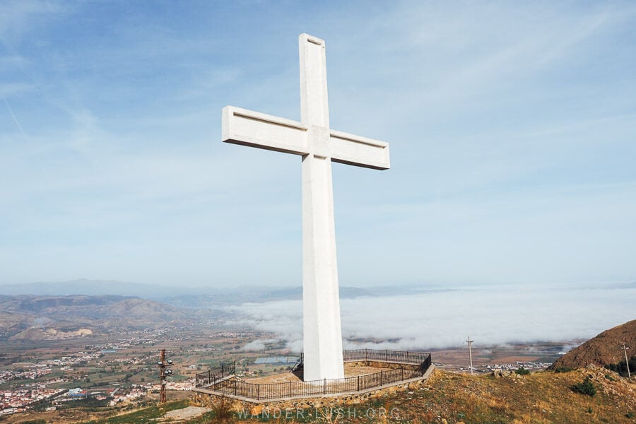 The Cross of Morava, a giant white cross above Korca, Albania, with a city panorama and low lying clouds.