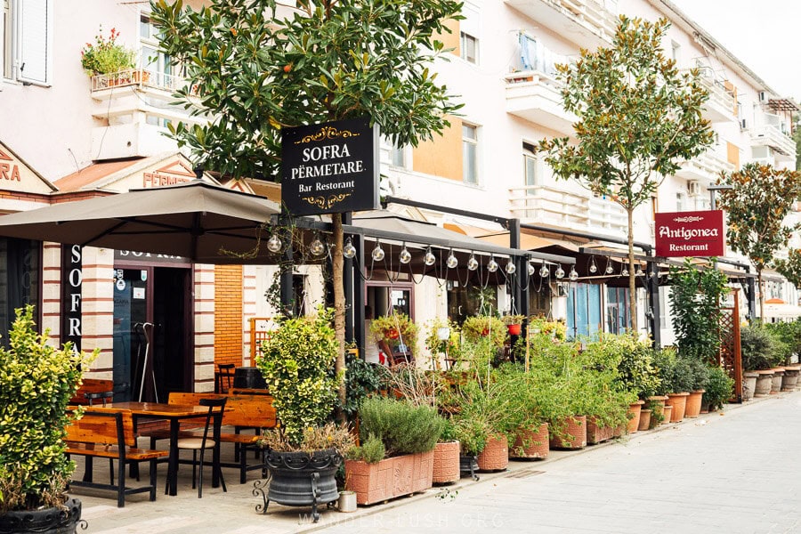 A street in Permet, Albania, with sidewalk restaurants and leafy plants.