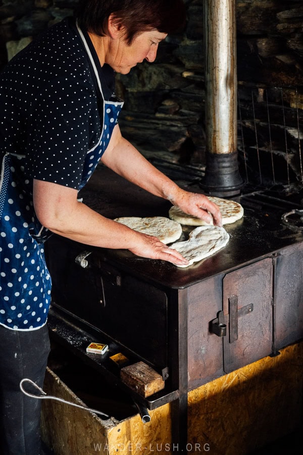 A woman in a blue apron prepares bread on a stove oven at a restaurant in Ushguli, Georgia.