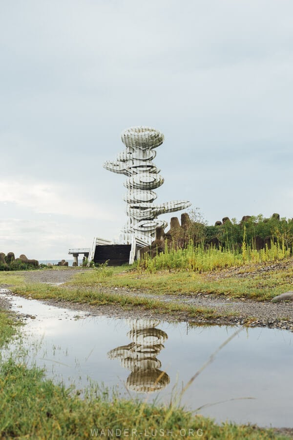 The Pier Sculpture in Anaklia viewed from a distance, with its mirror image seen in a puddle on the dirt road.