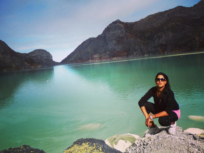 A woman wearing sunglasses crouches down on rocks before a green lake and mountain peaks.