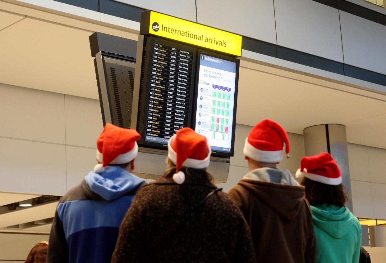 4 people wearing Santa hats at the airport are looking at the arrivals screen.