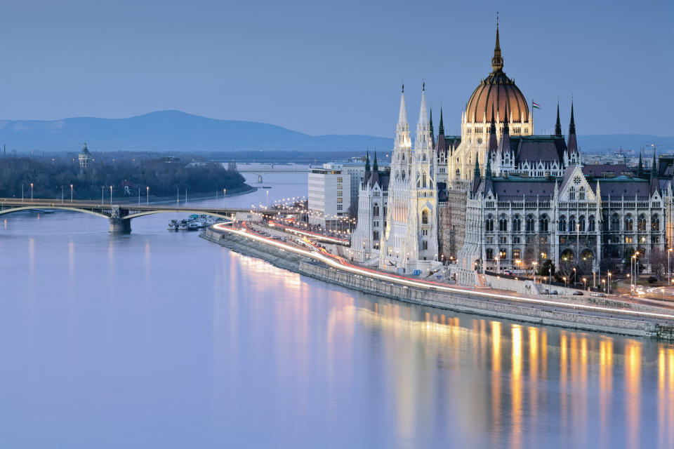 A serene view of the Hungarian Parliament Building alongside the Danube River at dusk, with faint lights reflecting on the water