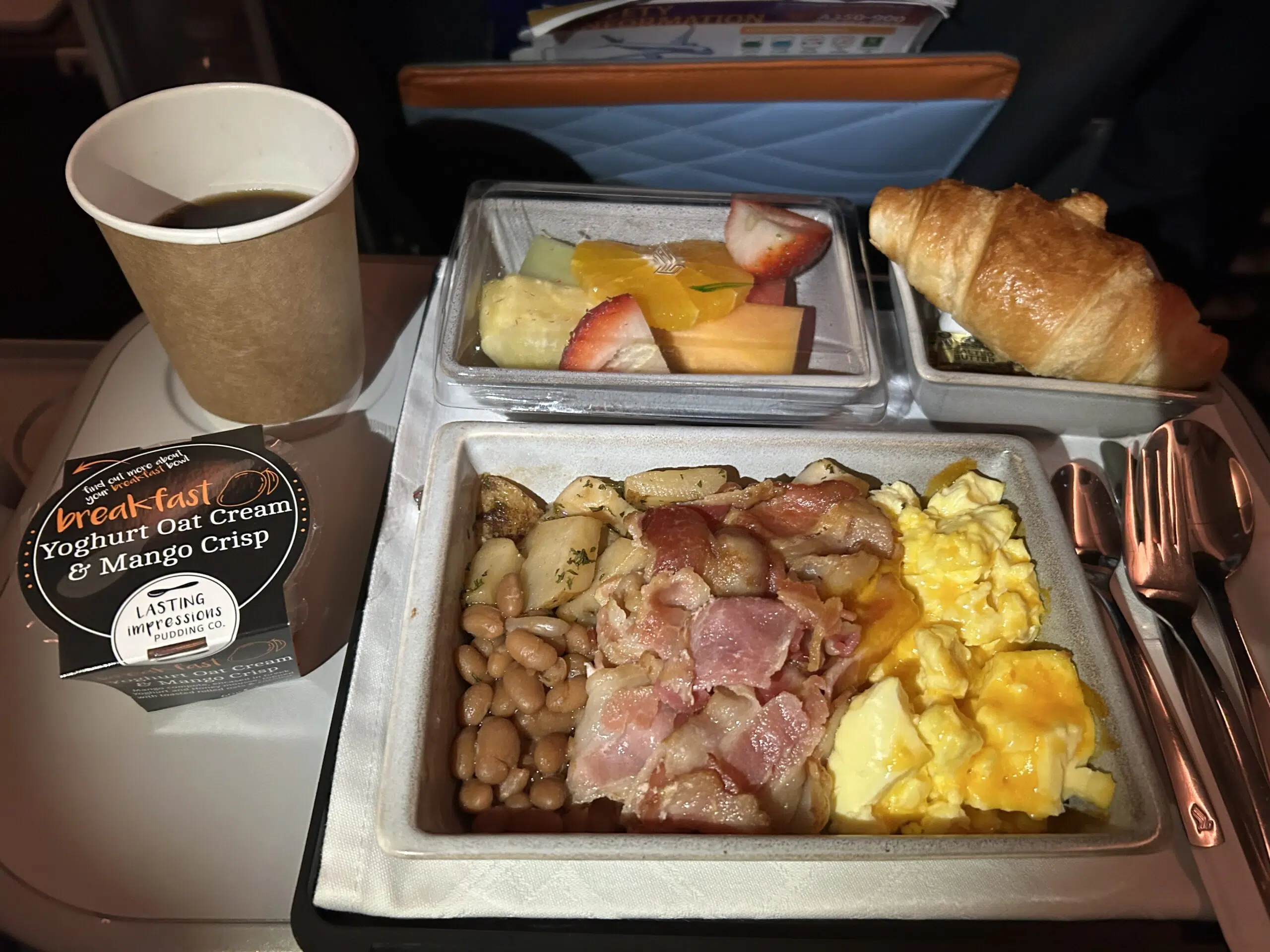 Eggs, bacon, beans, potatoes, fruit, bread, yogurt, and coffee on an airline tray