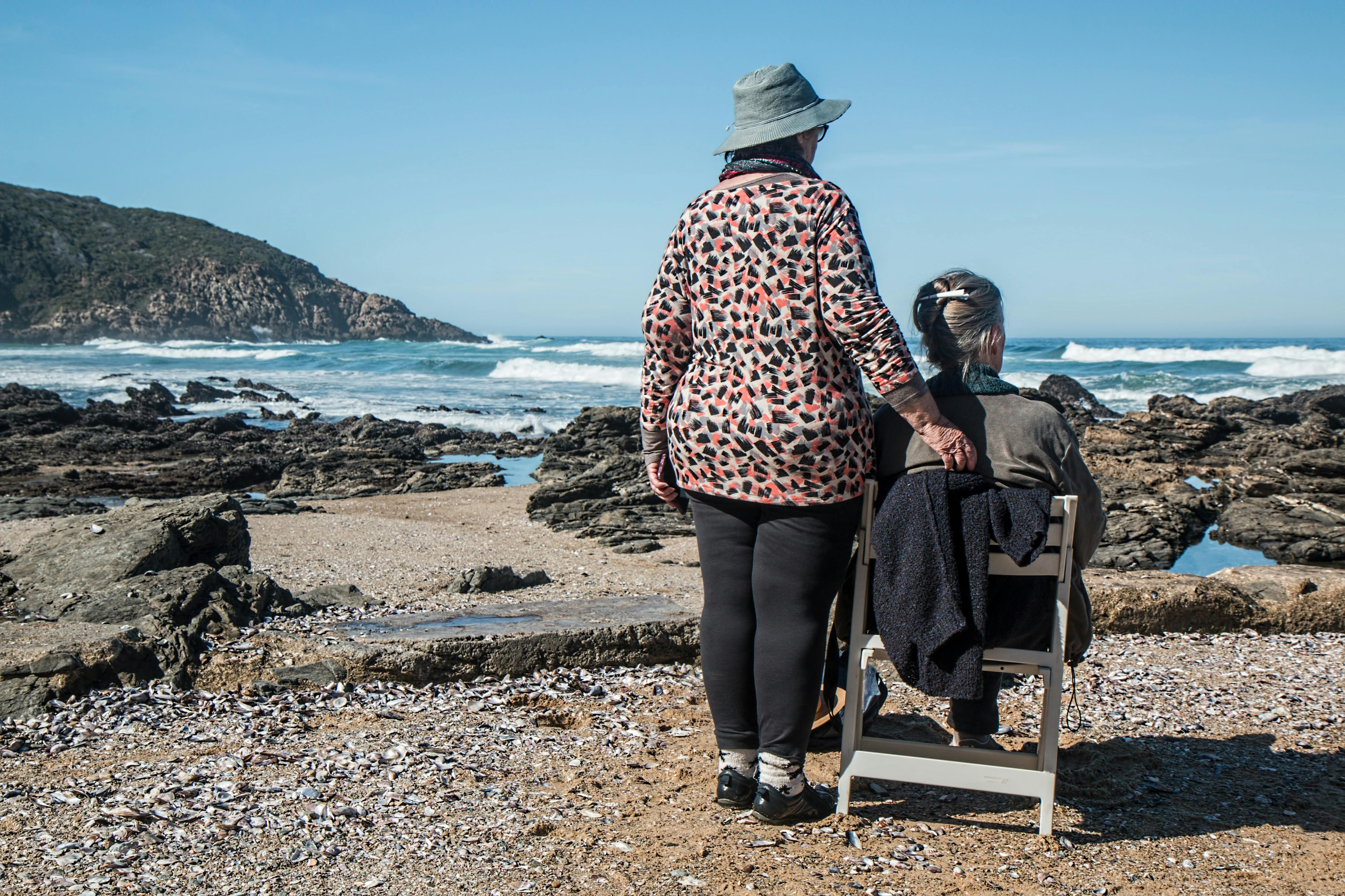 Two elderly women are at a rocky beach, one sitting in a chair while the other stands beside her, looking at the ocean.