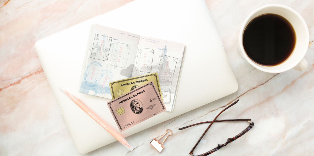 Two Amex Gold Card designs on top of table with an open passport, a pen, binder clip, glasses, and a cup of coffee