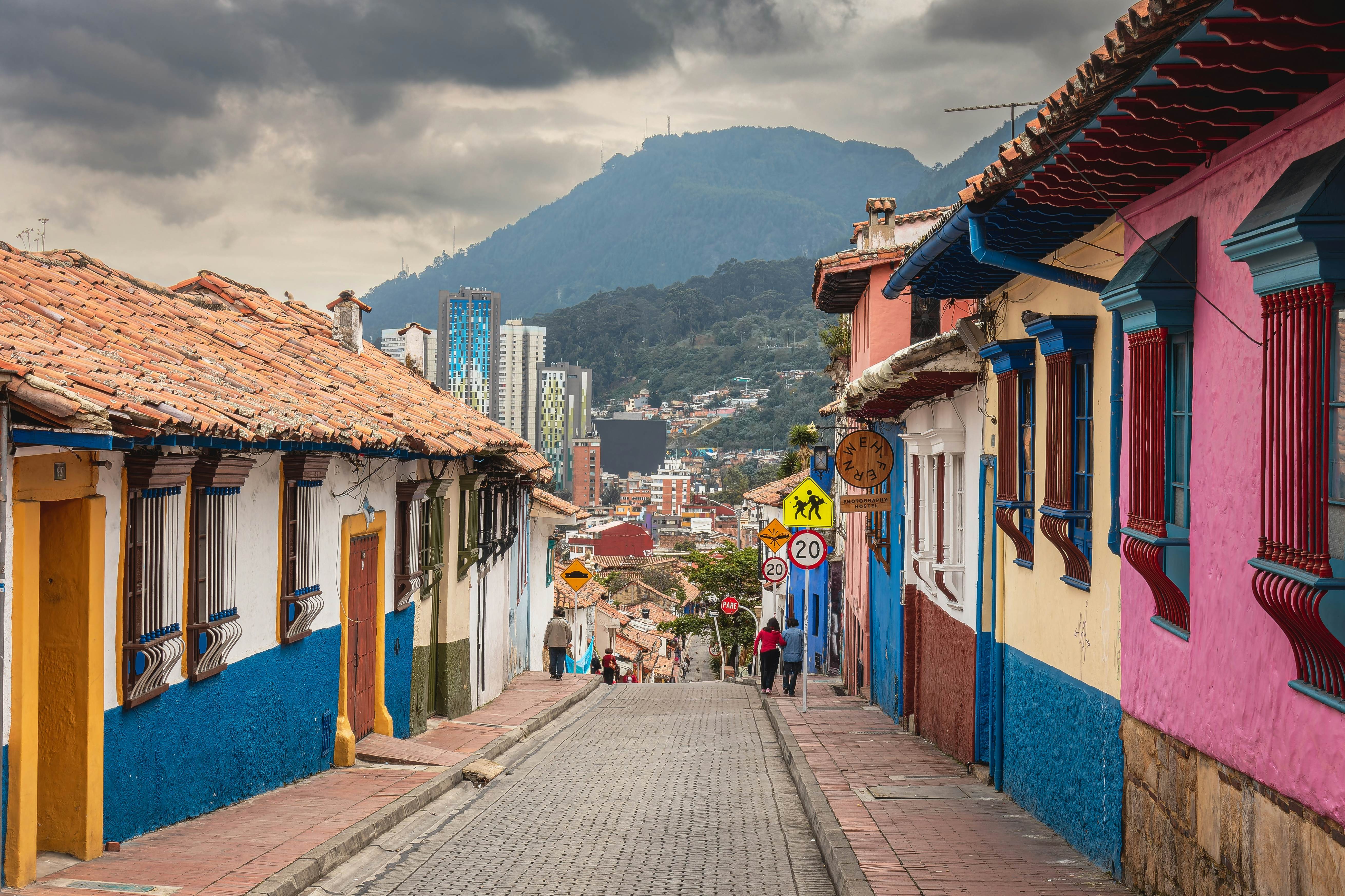 A narrow, cobbled street lined with one-story, brightly painted houses with cloudy mountains in the distance, La Candelaria, Bogotá, Colombia