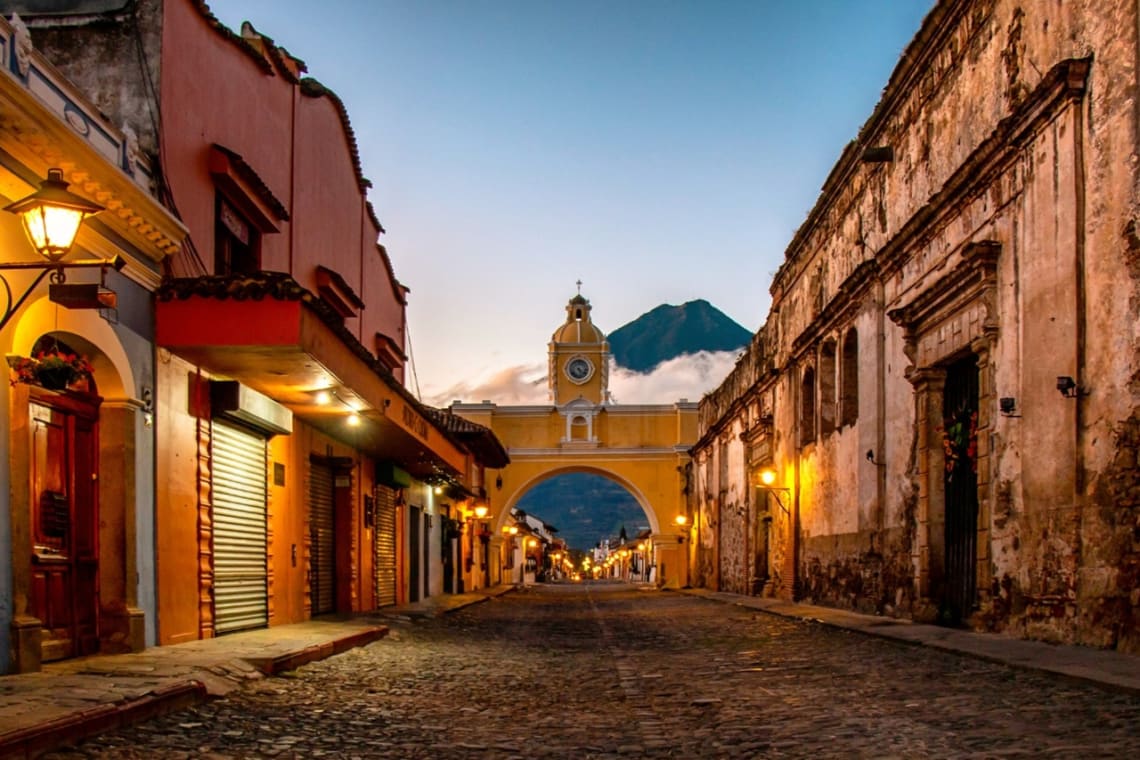 Cobblestone street with colonial buildings, yellow arch and volcano on the distance in Antigua