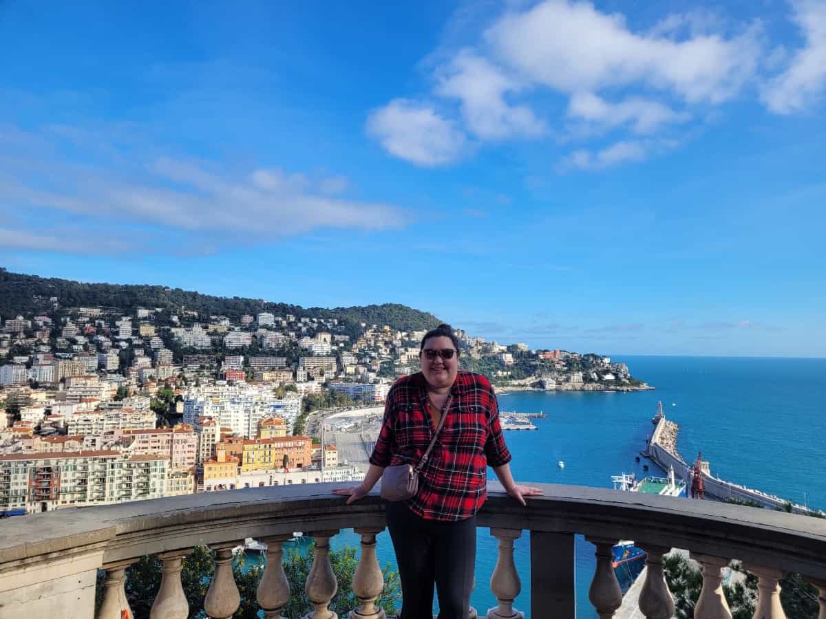 Riana leaning against a railing at the top of Nice Castle Hill with a view of the ocean, mountains and buildings behind her in Nice, France