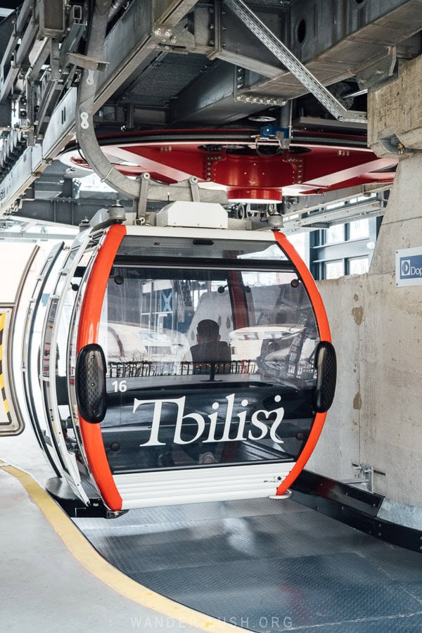 A modern cable car gondola with the name Tbilisi on it departs from a new lower station.