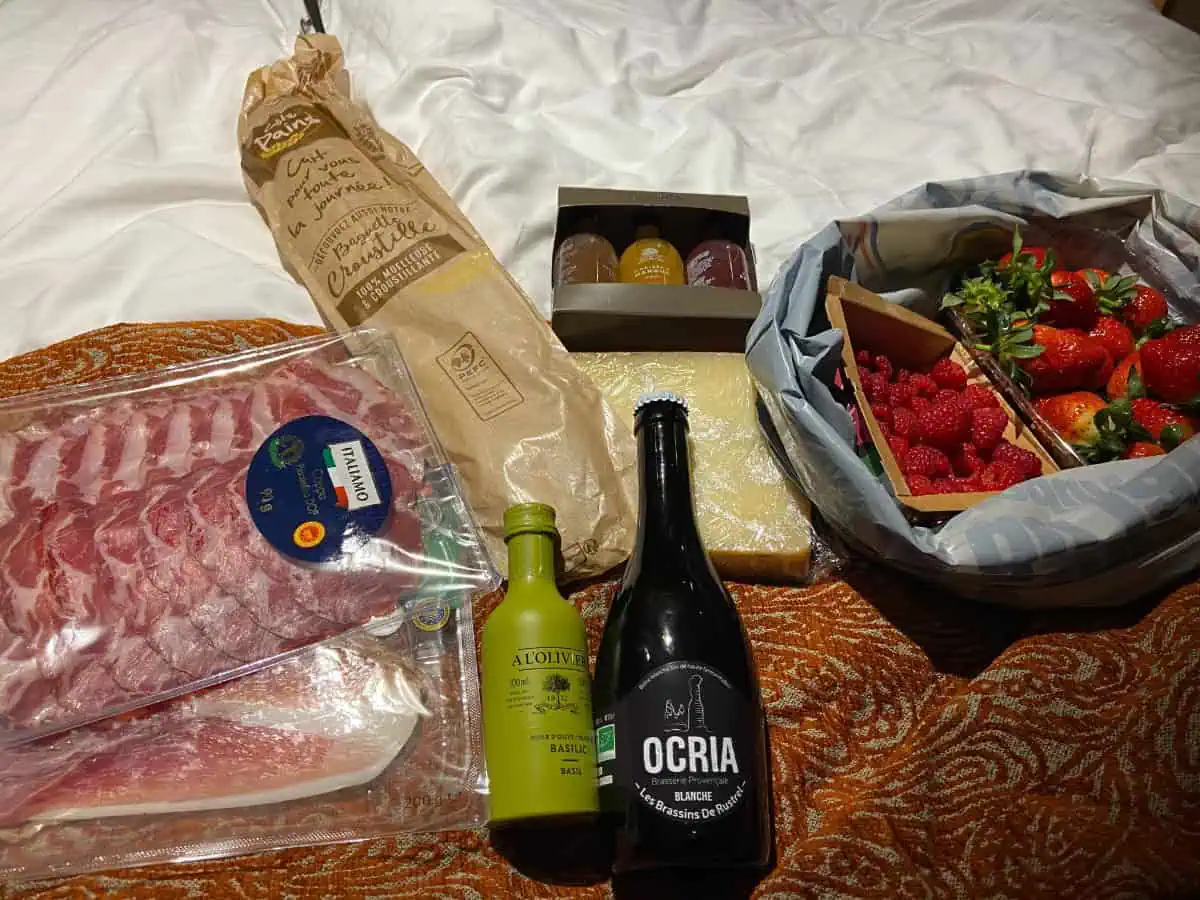 Picnic foods laid out on a bedspread including prosciutto, olive oil, baguette, baked goods, cheese and berries