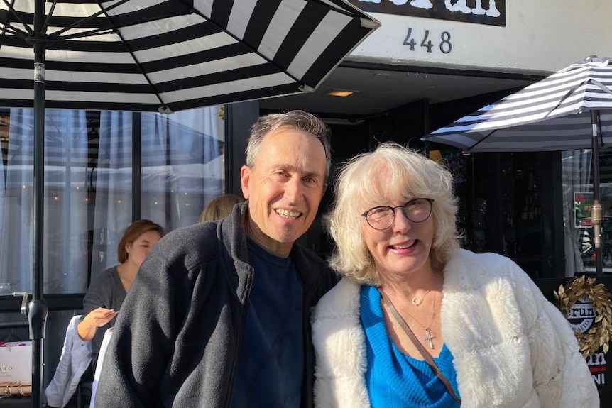 A man wearing a jumper and a woman wearing a white fur coat smile together in front of a cafe.