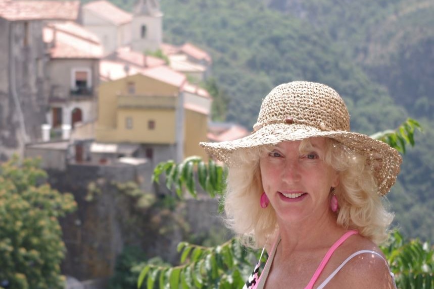 A woman smiles while wearing a hat and posing in front of a mountainous landscape.