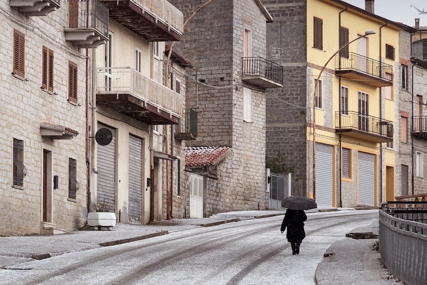 A person walks down an empty street lined with snow and bright buildings on either side.