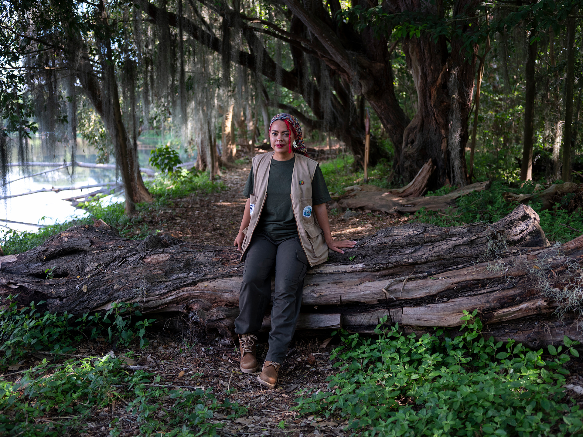 A woman with bright red hair sits on a fallen tree within a forest next to water