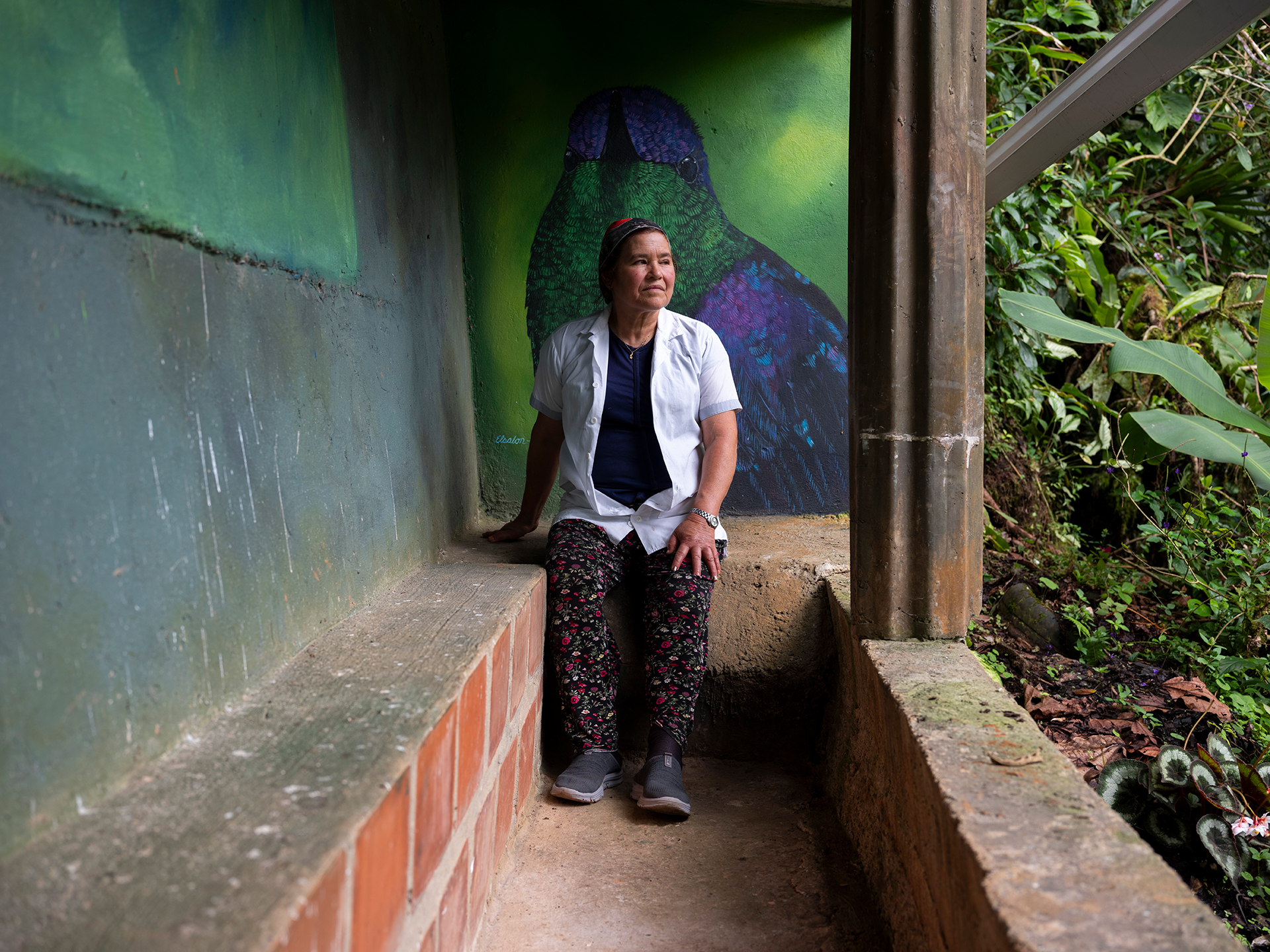 A woman sitting on a small brick wall with a green and purple painted bird mural behind her. She’s looking out at lush greenery to her left