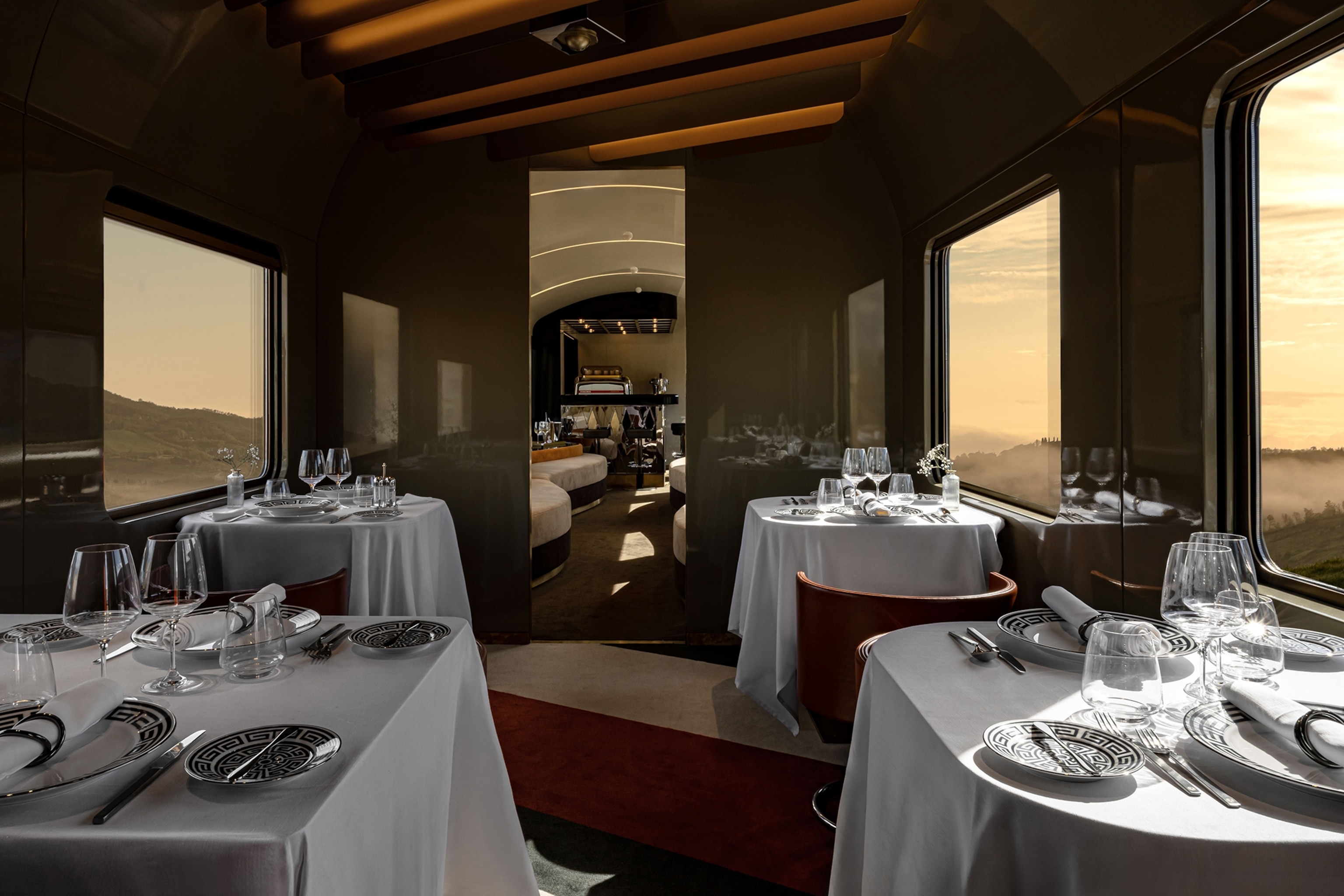 A view inside a train wagon with four tables set in white cloths and expensive silverware.